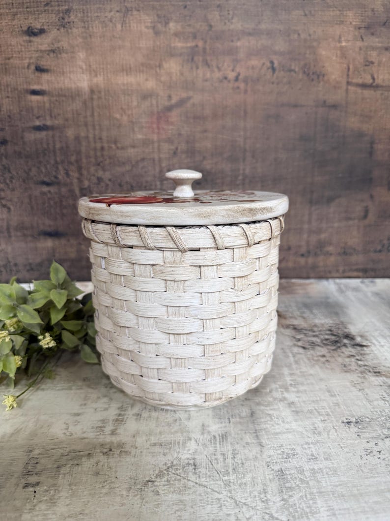 May include: A cream-colored woven basket with a lid. The lid has a small, round knob and a painted design. The basket is cylindrical and has a textured, woven appearance. The background is a rustic wooden surface.