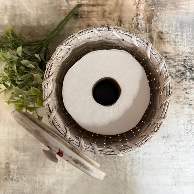 May include: A woven, off-white toilet paper holder with a lid, containing a roll of white toilet paper. The holder has a decorative woven pattern around the rim. A sprig of green leaves is visible in the background.