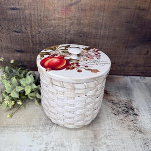 May include: A white woven basket with a lid featuring a red pomegranate illustration and botanical accents. The lid has a small white knob. The basket is set against a rustic wooden background, suggesting a farmhouse or vintage aesthetic.