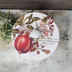 May include: A round, decorative lid with a white wooden knob. The lid features a botanical design with a large red pomegranate, berries, and floral elements. Text includes "DEBERN A PARIS" and a quote. The lid is likely for a storage container.