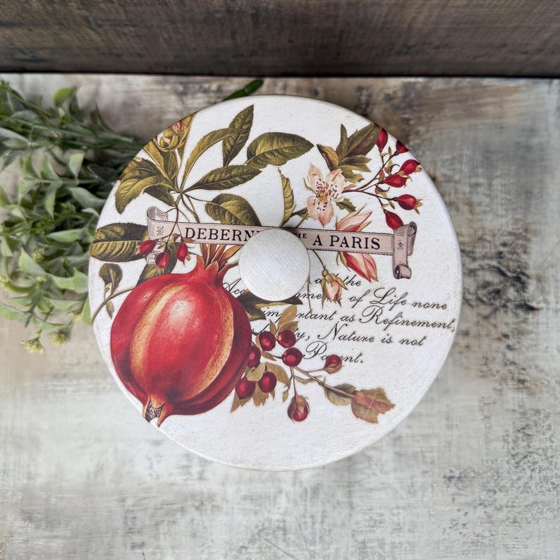 May include: A round, decorative lid with a white wooden knob. The lid features a botanical design with a large red pomegranate, berries, and floral elements. Text includes "DEBERN A PARIS" and a quote. The lid is likely for a storage container.