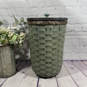 May include: A green woven basket with a lid and a green knob. The basket is sitting on a wooden floor.
