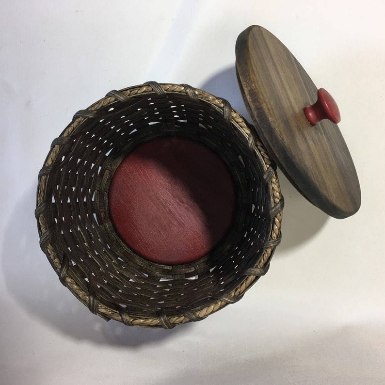 May include: A round woven basket with a dark brown and tan exterior. The basket has a red wooden lid with a small red knob. The lid is open and the inside of the basket is lined with a red fabric.