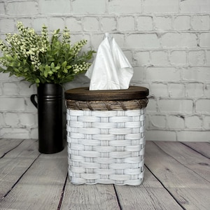 May include: A white woven tissue box cover with a dark brown wooden top. The tissue box is rectangular and has a woven pattern. A bundle of white tissues is coming out of the top.