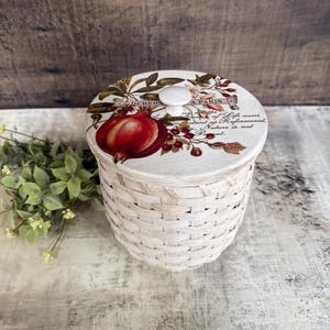 May include: A white woven basket with a lid featuring a pomegranate illustration and text. The lid has a small, round knob. The basket is cylindrical and sits on a light-colored surface. The text on the lid reads "DEHERS A PARIS".