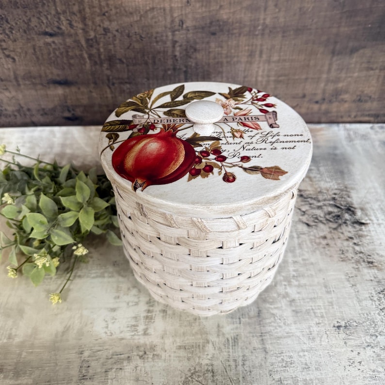 May include: A white woven basket with a lid featuring a pomegranate illustration and text. The lid has a small, round knob. The basket is cylindrical and sits on a light-colored surface. The text on the lid reads "DEHERS A PARIS".