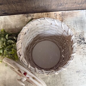 May include: A woven basket with a matching lid. The basket is light beige with a round base and a woven design. The lid is decorated with a floral pattern and a small handle. The basket is empty.