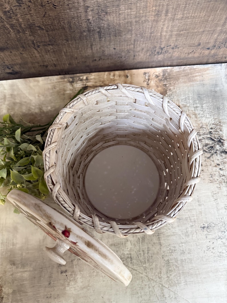 May include: A woven basket with a matching lid. The basket is light beige with a round base and a woven design. The lid is decorated with a floral pattern and a small handle. The basket is empty.