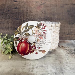May include: A decorative round lid with a white knob, featuring a red pomegranate illustration and botanical accents. The lid is next to a woven basket. Text on the lid reads "DEBER...A PARIS...of Life...Nature is not...Pant."
