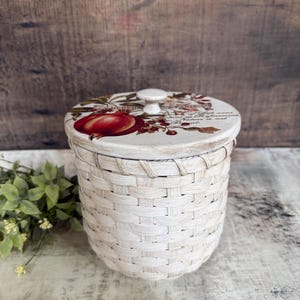 May include: A white woven basket with a decorative lid. The lid features a red fruit illustration and floral accents. The basket is cylindrical and has a small white knob on top. The basket is approximately 6 inches tall.
