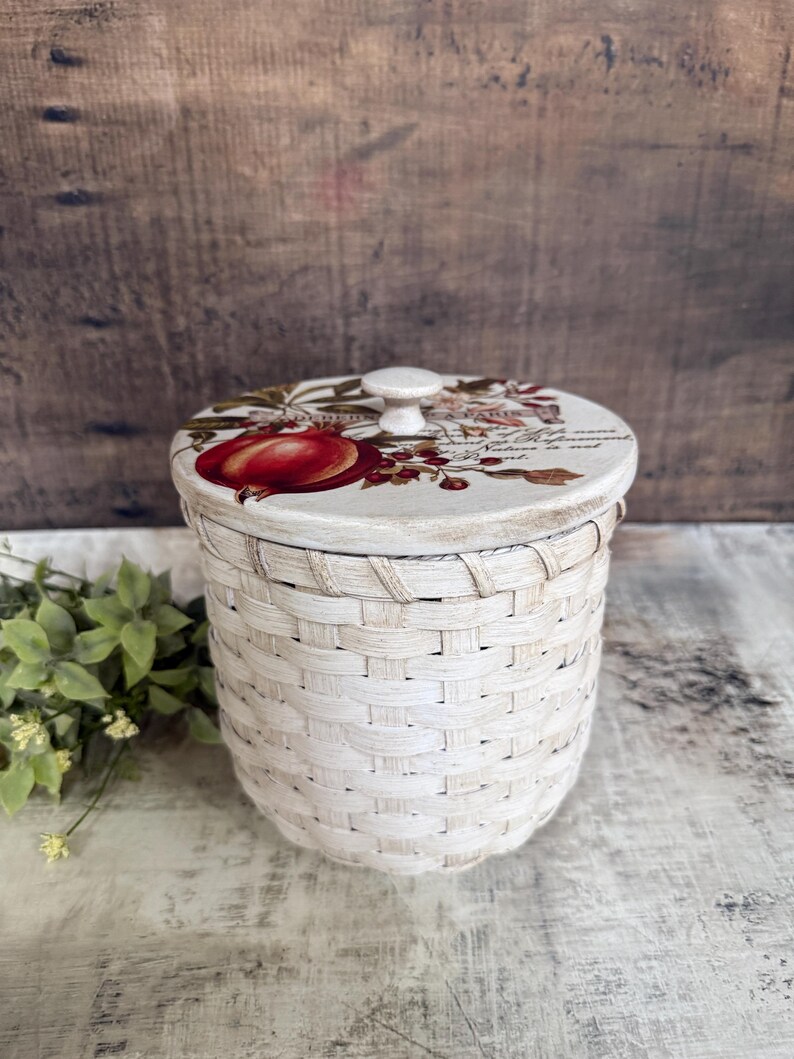 May include: A white woven basket with a decorative lid. The lid features a red fruit illustration and floral accents. The basket is cylindrical and has a small white knob on top. The basket is approximately 6 inches tall.