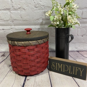 May include: A red woven basket with a wooden lid and a red knob. The basket is sitting on a wooden floor with a black sign that says "SIMPLIFY" in gold lettering.
