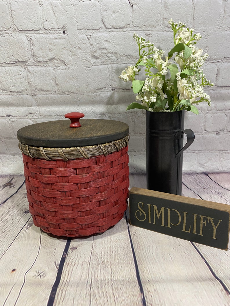 May include: A red woven basket with a wooden lid and a red knob. The basket is sitting on a wooden floor with a black sign that says "SIMPLIFY" in gold lettering.