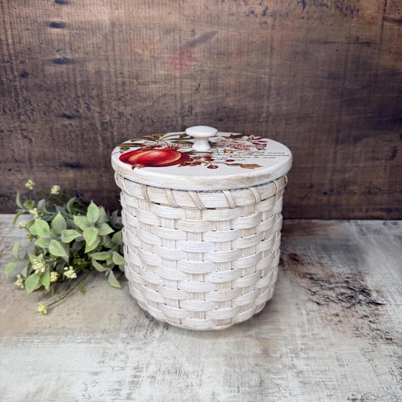 May include: A white woven basket with a decorative lid. The lid features a red apple design and a small white knob. The basket is cylindrical and has a rustic, farmhouse aesthetic. The basket is approximately 6 inches in diameter and 6 inches tall.