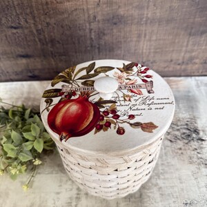 May include: A round, woven basket with a decorative lid. The lid features a botanical design with a large red pomegranate, berries, and text that reads "A PARIS". The basket is white with a natural woven texture.