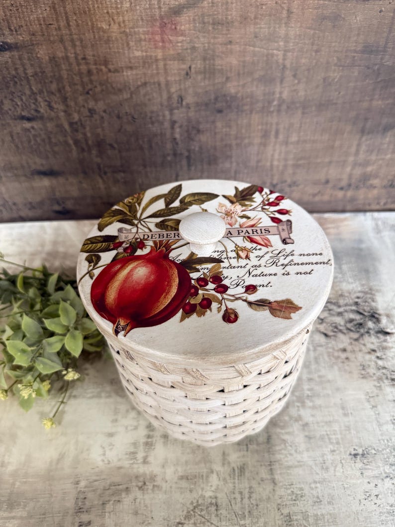May include: A round, woven basket with a decorative lid. The lid features a botanical design with a large red pomegranate, berries, and text that reads "A PARIS". The basket is white with a natural woven texture.