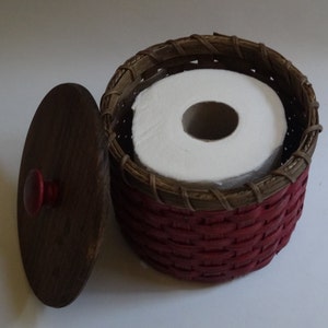 May include: A red and brown woven basket with a lid, containing a roll of white toilet paper. The lid has a red knob.