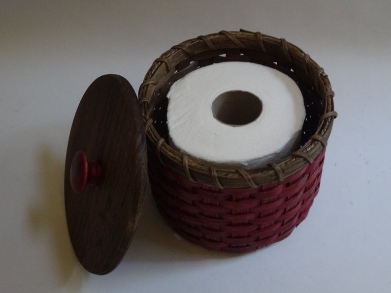 May include: A red and brown woven basket with a lid, containing a roll of white toilet paper. The lid has a red knob.