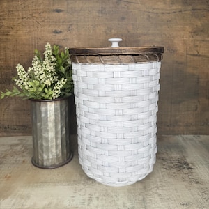 May include: A cylindrical woven basket with a white finish, featuring a dark brown wooden lid and a small white knob handle. The basket has a tight, uniform weave pattern throughout its body and a decorative braided trim along the top edge.