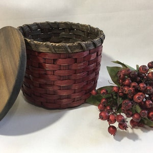 May include: A red woven basket with a wooden lid and a red knob. The basket is sitting on a white surface next to a sprig of red berries.