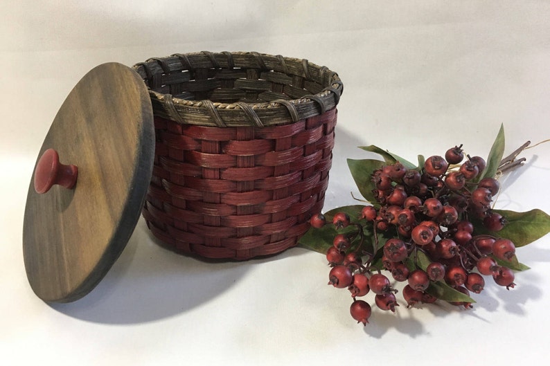 May include: A red woven basket with a wooden lid and a red knob. The basket is sitting on a white surface next to a sprig of red berries.