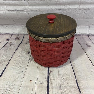 May include: A red woven basket with a wooden lid and a red knob. The basket is sitting on a wooden surface.