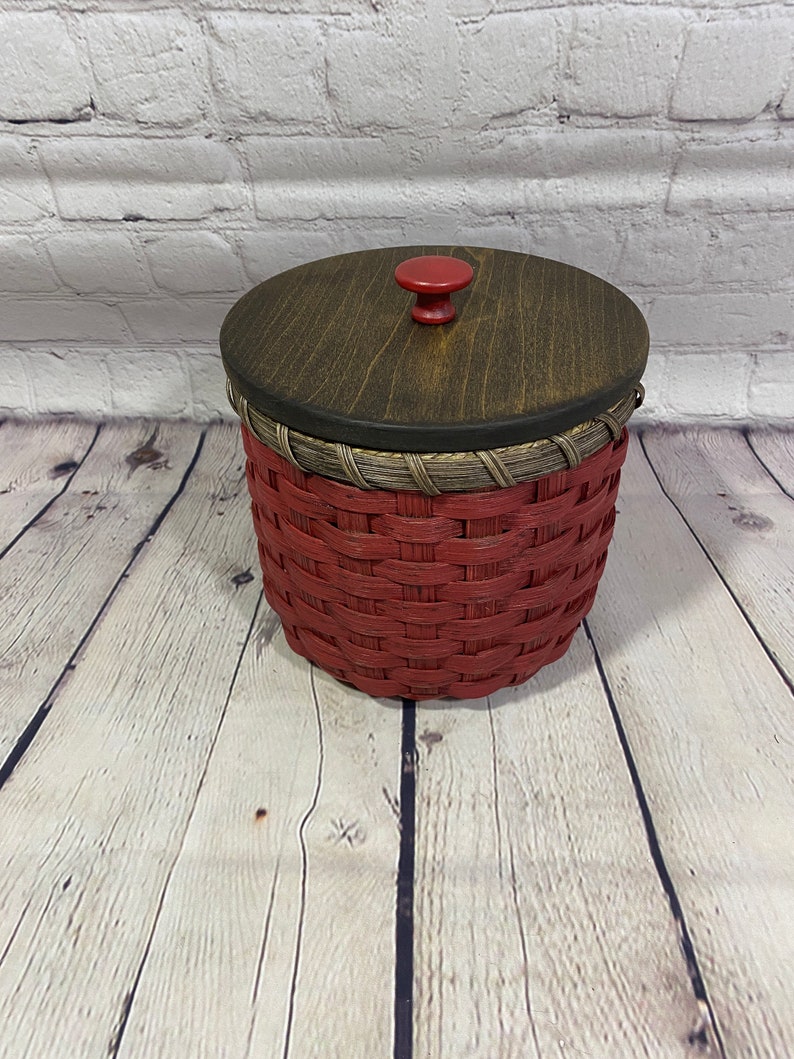 May include: A red woven basket with a wooden lid and a red knob. The basket is sitting on a wooden surface.
