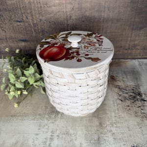 May include: A white woven basket with a lid featuring a pomegranate design. The lid has a white knob and text that reads "DEBERS A PARIS". The basket is decorated with a botanical design and is suitable for storage.