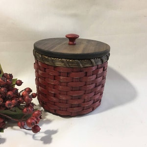 May include: A red woven basket with a wooden lid and a red knob. The basket has a brown and gold trim around the top.