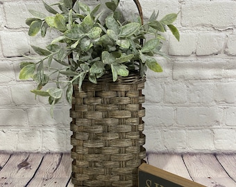 May include: A brown woven basket with a handle, filled with green leafy plants, sits on a wooden surface with a black sign that reads "SIMPLIFY".