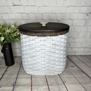May include: A white woven basket with a dark brown wooden lid and a small white knob. The basket is cylindrical and sits on a wooden surface. The background is a white brick wall.