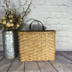 May include: A woven wicker basket with a brown leather handle. The basket is square shaped and has a light brown color.