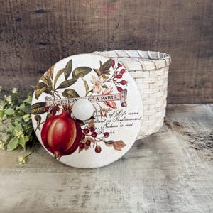 May include: A decorative round lid with a white knob, featuring a red pomegranate illustration and text that reads "DEBERN C A PARIS". The lid is next to a woven basket, with a wooden background.