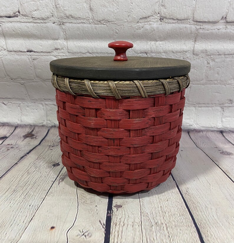 May include: A red woven basket with a wooden lid and a red knob. The basket has a brown woven trim around the top.