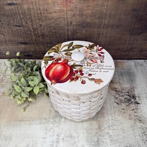 May include: A white woven basket with a lid featuring a pomegranate design. The lid has a white knob and botanical illustrations. The text on the lid reads "DEBER A PARIS" and a quote. The basket is on a light-colored surface.