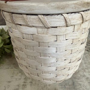 May include: A white woven basket with a lid. The lid is decorated with a floral design in shades of red and brown. The basket has a handle on the lid and is made of a natural material.