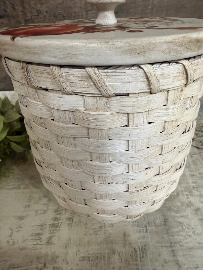 May include: A white woven basket with a lid. The lid is decorated with a floral design in shades of red and brown. The basket has a handle on the lid and is made of a natural material.