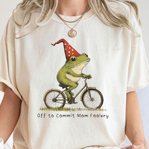 May include: Cream-coloured t-shirt with a playful design of a green frog on a bicycle, wearing a red and white pointed hat. The text "Off to Commit Mom Foolery" is printed beneath the image. The shirt has short sleeves and a relaxed fit.