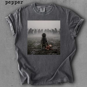May include: Dark gray t-shirt with a printed image of a child holding a teddy bear in a field with a crowd of people in the background. The word "pepper" is printed in the upper left corner.