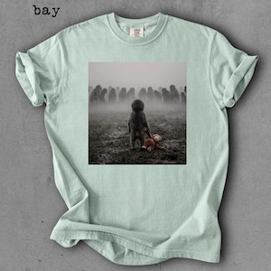 May include: A light green t-shirt with a square graphic of a child holding a teddy bear in a foggy field. The word "bay" is printed in the upper left corner of the shirt.