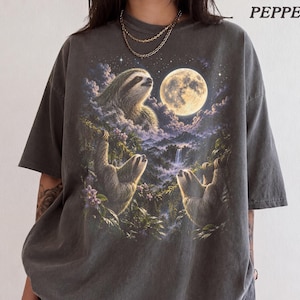 May include: A charcoal grey t-shirt featuring a graphic print of three sloths in a lush jungle landscape under a large full moon and starry night sky. The word PEPPER is printed in the upper right corner of the shirt in a simple font.