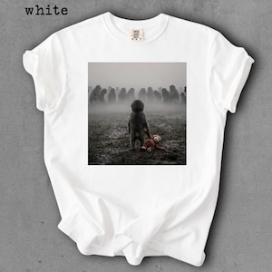 May include: White t-shirt with a square graphic featuring a child figure holding a teddy bear in a misty field, with a crowd of indistinct figures in the background. The word "white" is printed at the top left.