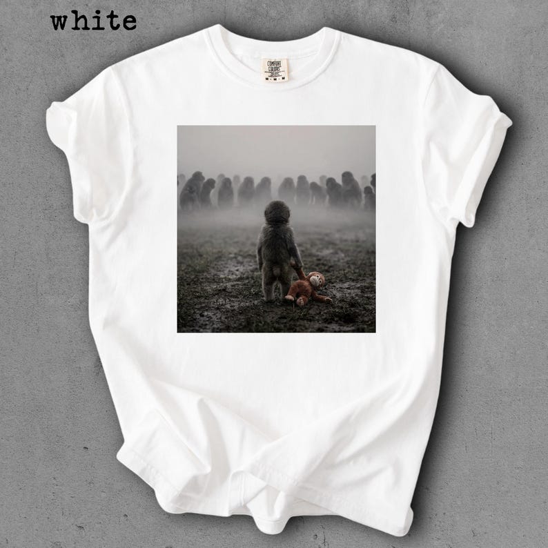 May include: White t-shirt with a square graphic featuring a child figure holding a teddy bear in a misty field, with a crowd of indistinct figures in the background. The word "white" is printed at the top left.