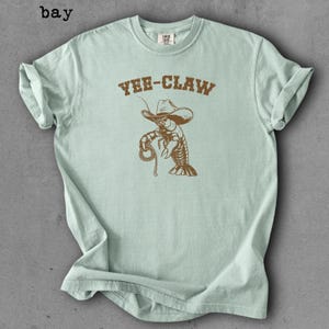 May include: Light blue t-shirt with a brown graphic of a cowboy lobster holding a rope, and the text "YEE-CLAW" above it. The shirt has a crew neck and short sleeves. The word "bay" is printed in the upper left corner.