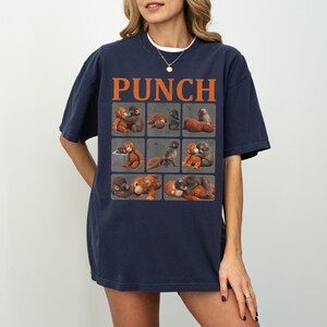 Peut inclure: T-shirt bleu marine avec le mot "PUNCH" en lettres orange. Le t-shirt pr&eacute;sente une grille de neuf carr&eacute;s, chacun contenant une illustration &agrave; l'aquarelle de singes dans diverses poses. Le t-shirt est de coupe d&eacute;contract&eacute;e.