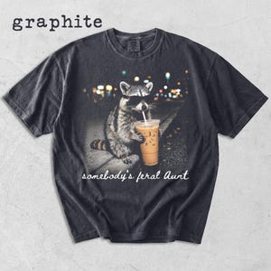 May include: A graphite-colored short-sleeve t-shirt featuring a graphic of a raccoon wearing sunglasses and holding an iced coffee. Below the raccoon, the text reads "somebody's feral Aunt" in a white script font. The shirt has a crew neckline.