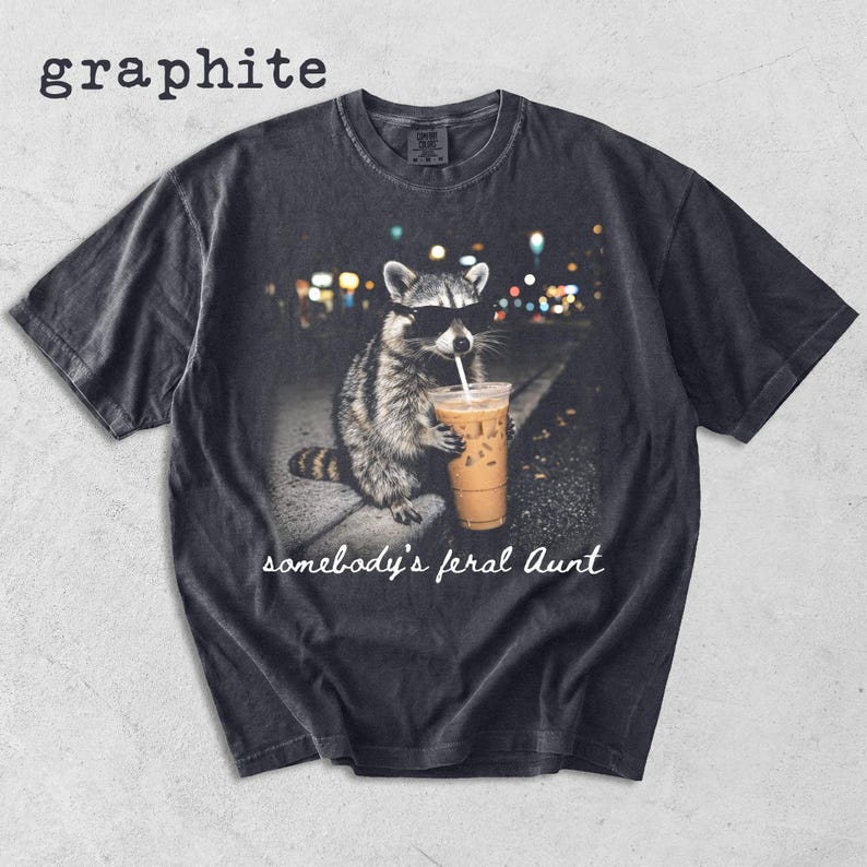 May include: A graphite-colored short-sleeve t-shirt featuring a graphic of a raccoon wearing sunglasses and holding an iced coffee. Below the raccoon, the text reads "somebody's feral Aunt" in a white script font. The shirt has a crew neckline.