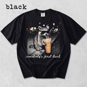 May include: A black short-sleeve t-shirt featuring a graphic of a raccoon wearing black sunglasses and holding an iced coffee with a straw. Below the raccoon, the text reads "somebody's feral Aunt" in a white cursive script font.