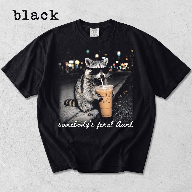 May include: A black short-sleeve t-shirt featuring a graphic of a raccoon wearing black sunglasses and holding an iced coffee with a straw. Below the raccoon, the text reads "somebody's feral Aunt" in a white cursive script font.