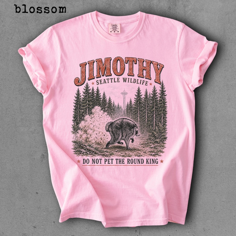 Op de afbeelding: Een lichtroze T-shirt met korte mouwen en een afbeelding van een ronde wasbeer in een bos met de Space Needle op de achtergrond. De tekst luidt JIMOTHY SEATTLE WILDLIFE en DO NOT PET THE ROUND KING in oranje en zwarte letters.